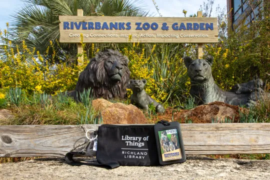 A Riverbanks Zoo and Garden entrance sign stands behind bronze lion statues surrounded by greenery. In the foreground, a Richland Library “Library of Things” pouch and a Riverbanks Zoo and Garden community pass rest on a wooden ledge.
