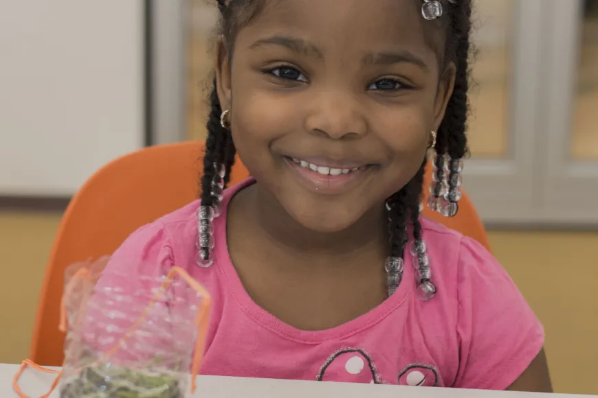 Young black female with a bright bow in her hair and a pink shirt sits in front of a table covered with newspaper during a gardening program at Richland Library