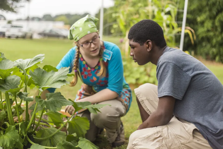 White staff member in bright blue shirt and green head scarf talks with black teen male next to a garden bed at Richland Library