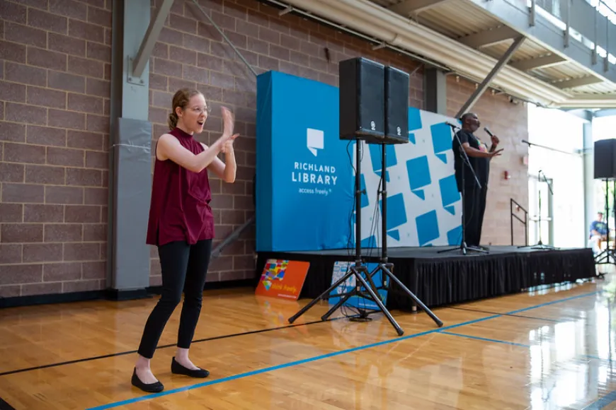 A sign language interpreter stands in the foreground, actively interpreting, while a speaker presents on a small stage behind her at Richland Library.