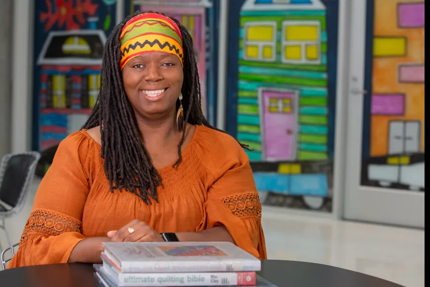 A smiling person sits at a table with a stack of books, wearing an orange top and a colorful headwrap, with bright, illustrated house artwork in the background.