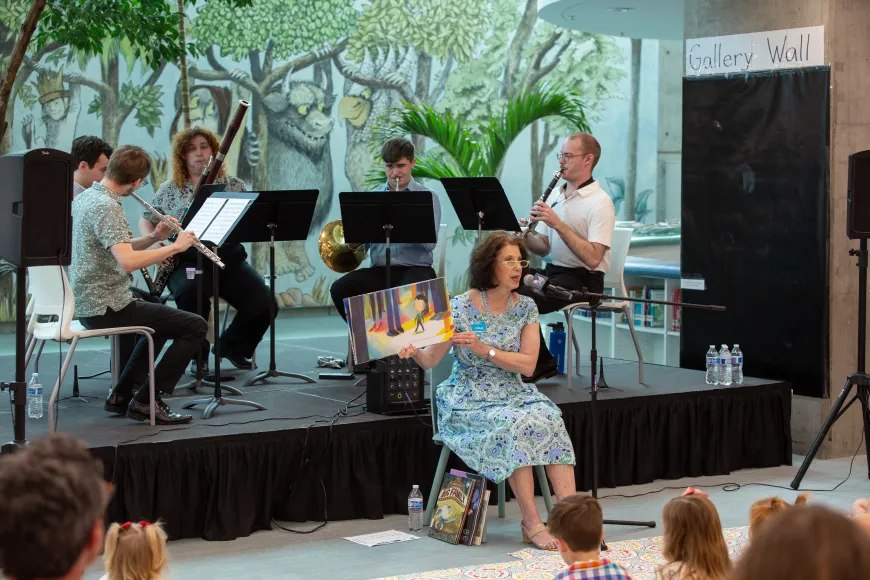 Small musical ensemble performs behind a storyteller reading a picture book to children seated on the floor.