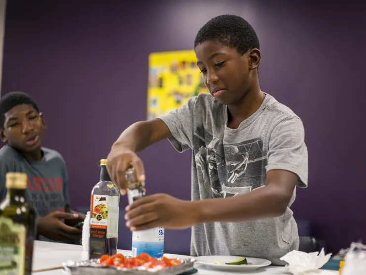 Black male wearing a grey tshirt standing in front of a purple wall adds seasoning to sliced tomatoes
