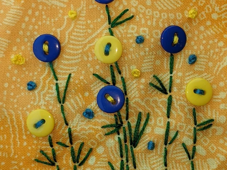 An image of cheerfully bright yellow patterned fabric with flowers embroidered on using blue and yellow buttons and green thread for stems and leaves