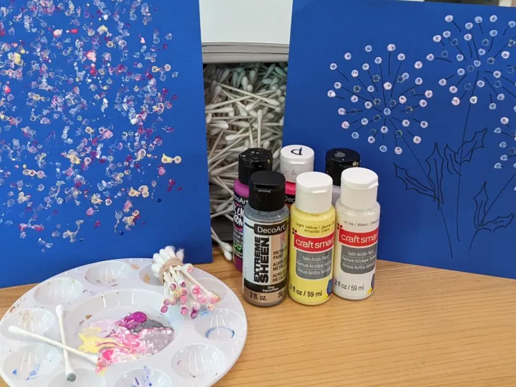 Photo: two dark blue pieces of cardstock painted with abstract designs, along with a bin of cotton swabs, several bottles of acrylic paint in various colors, a white paint palette, and a bundle of cotton swabs with paint on them