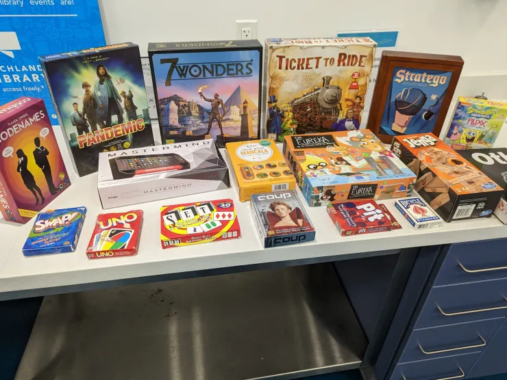 Image of a group of various board and card games arranged on a white table with a white wall in the background. Board games include Pandemic, 7 Wonders, Pit, Coup, etc.