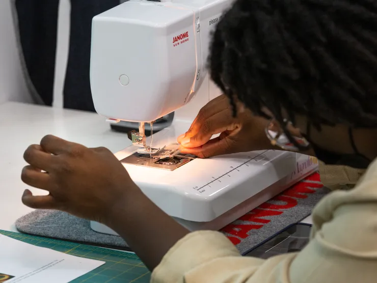 Female with black hair and tan shirt pushes fabric through a sewing machine at Richland Library