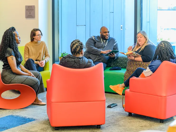 Six adults participate in a book club. They sit on orange-red chairs.
