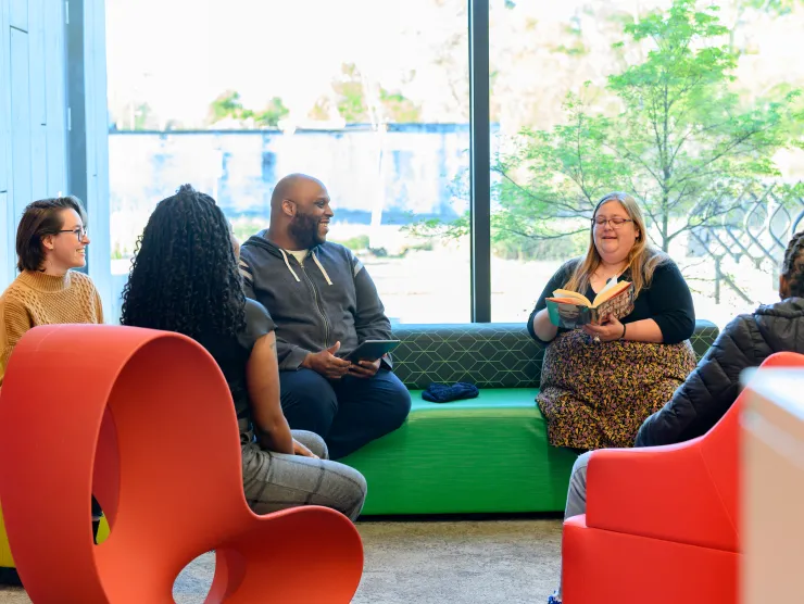 Six adults participate in a book club. They sit on orange-red chairs.