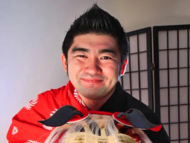 mage of Yasu Ishida, an Asian man with dark hair styled to a point dressed in a red traditional garment holding a red and black Asian dragon mask.