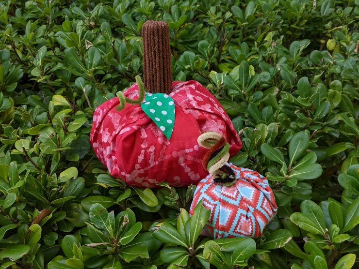 Two fabric pumpkins, one large one that's red with a straight brown yarn stem and one small one that's red and blue with chenille sticks as a stem