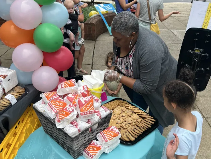 Woman in grey sweater giving out popcorn and cookies beside a balloon arch.