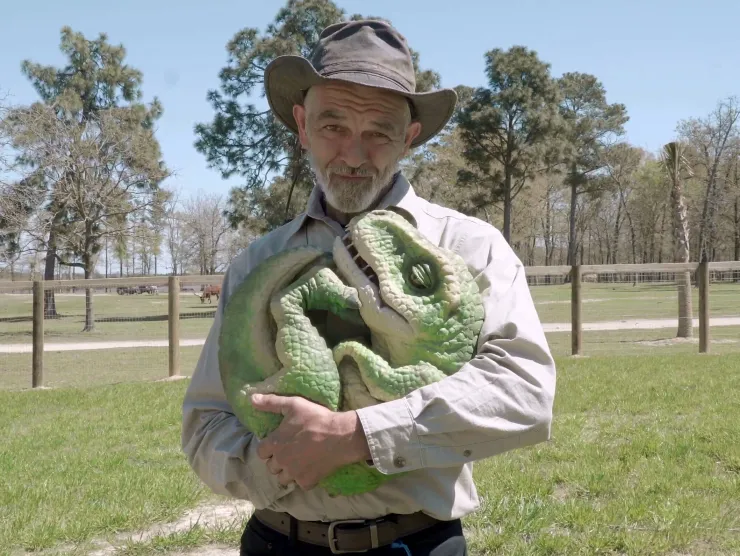 Image of Mr. Ed, an older white man.  He wears a hat and safari-style clothing.  In his arms, he cradles a dinosaur puppet.