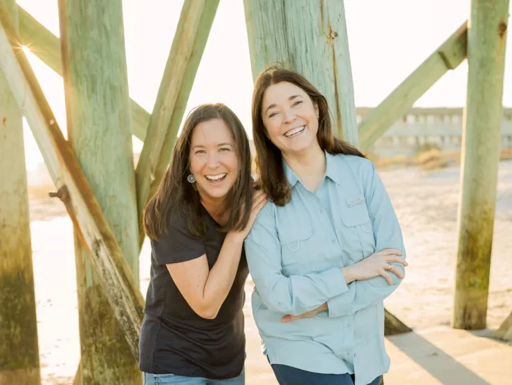 Photo of Mary Alice Monroe and Angela May.  Two white women are standing underneath a pier on a beach.  The woman on the left wears a dark t-shirt, jeans and sandals.  Her hands are on the shoulders of the other woman and she is smiling.  The woman on the right is wearing a light denim long-sleeved button-down shirt with dark jeans and sandals.  Her arms are crossed and she is also smiling.  
