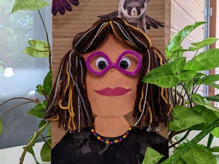 A cartoonish self portrait of a female with purple glasses, brown and grey hair, black shirt, and a rainbow necklace with a kestrel flying over her shoulder