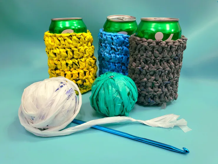 can koozies in yellow blue and grey crocheted plastic yarn. two small balls of plastic yarn in white and green with a blue crochet hook