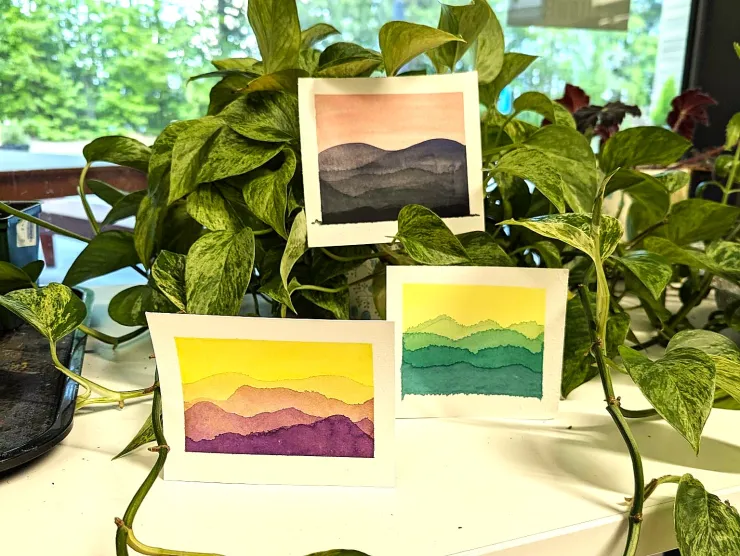 Three mini-watercolors of mountain landscapes. One is of black mountains with a pink sky. One is purple mountains with a yellow sky. And the other is green mountains with a yellow sky.