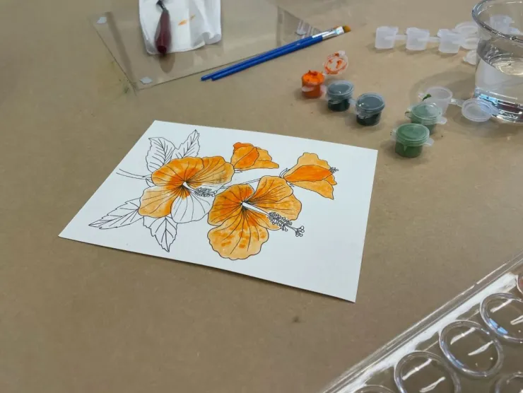 Water color paper that is printed with a flower. The flower petals are painted in orange water colors. Next to the paper is a paint brush and a few small painter containers.