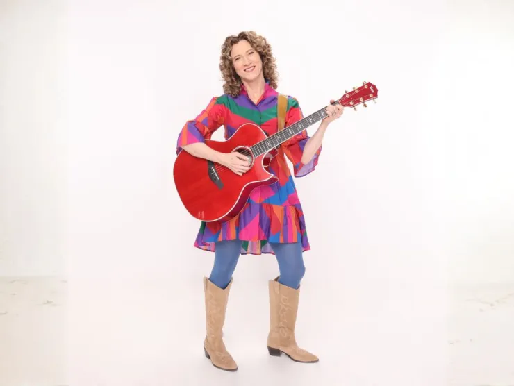 A woman  in cowboy boots and a colorful dress holding an acoustic guitar.