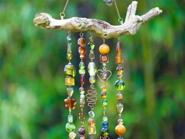 A multi-colored beaded wind chime hanging from a dry branch.  The is blurred green foliage in the background.