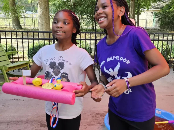 Two African American tweens holding up their found materials boat made of pink pool noodles , cardboard, and wooden dowels.