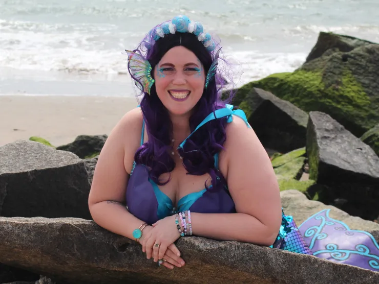 Image of Buoy the Brooklyn Mermaid.  She is a white woman on a beach perched on a rock.  She has a white and turquoise seashell headband and iridescent gills attached to her face.  She wears colorful make-up in blues and purples and a mermaid tail.  She is smiling.  