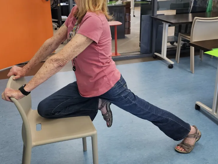 Women with left knee on chair and right leg extended and straight back. Arms are out straight and holding the to of the chair.