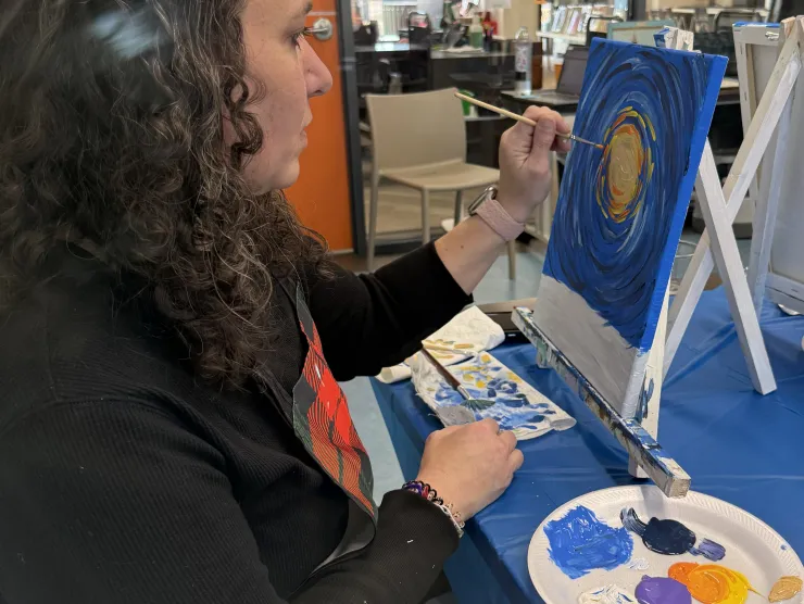 Women Painting in canvas in round pattern with the center being yellow and continuing with white orange, purple, blue, black, with the background being a bright blue. 