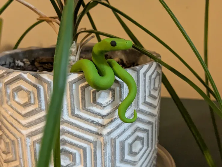 A small green snake made of polymer clay rests on the edge of a grey and white pot holding an ornamental grass plant.