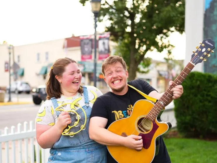 Photo of the BenAnna Band.  On the left, Anna is a white woman wearing denim overall shorts and a pale yellow banana t-shirt.  She's looking at Ben and holding a banana-shaped tambourine.  Ben is on the right.  He is strumming a guitar while wearing a black BenAnna Band t-shirt and navy shorts.  Both Ben and Anna are smiling.  
