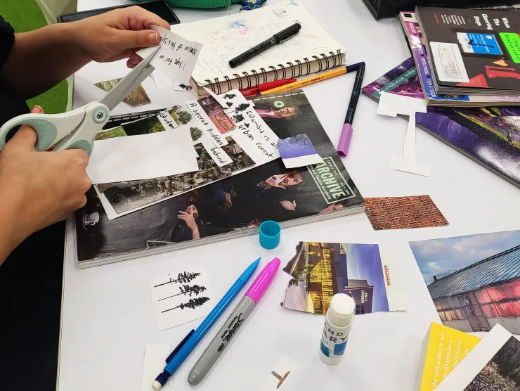 person cutting text from paper to glue onto collage table with glue stick pens and magazines