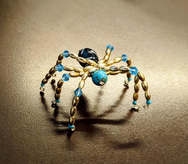 Beaded spider with a turquoise head, black body, and gold rice beads and blue crystals as legs.
