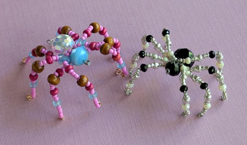 Two beaded spiders. One is pink, light blue, magenta, and brown. One is silver and black.