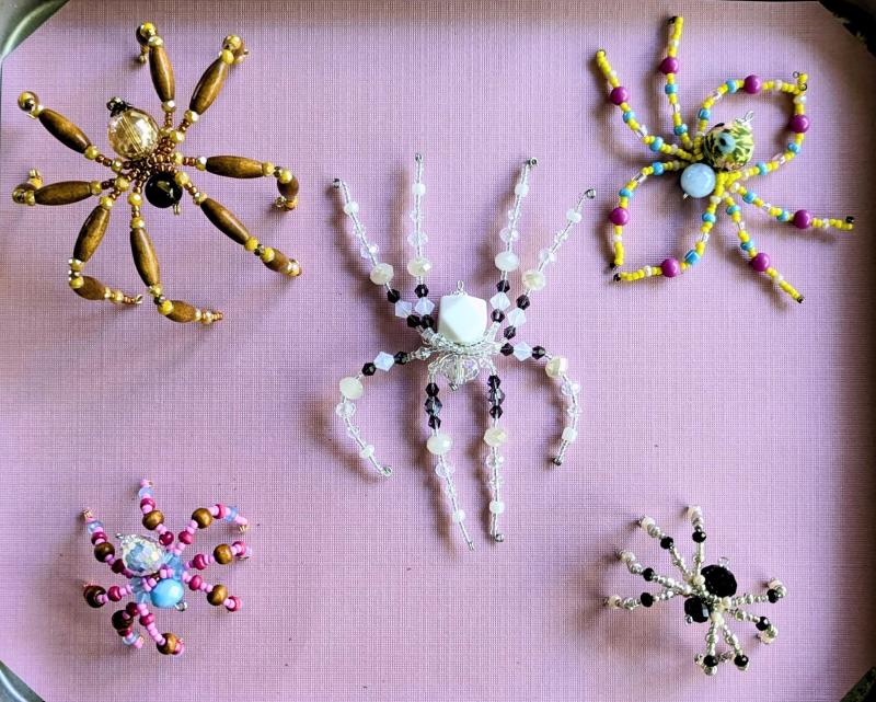 A tray of 5 beaded spiders. One is white and purple. One is yellow, blue, and pink. One is brown wood with tiger's eye stone and gold beads. One is silver and black. One is pink, brown, blue, and magenta.