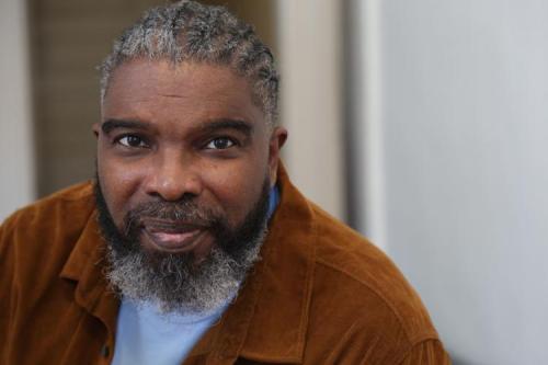Photo of Darion McCloud.  Darion is an older Black man.  He has braided salt-and-pepper hair and a beard.  He wears a brown jacket and light blue t-shirt.  He has a friendly expression on his face.  