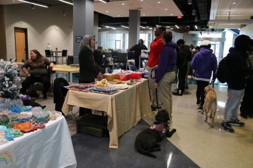 Market Vendor in grey sweater speaking with a person in a red jacket and a person in a purple hoodie. There are also other customers standing and walking around speaking with other market vendors and service dogs are pictured.