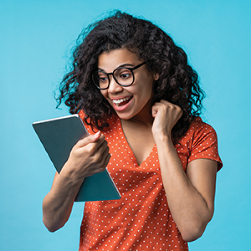 Teenage girl wearing a bright orange shirt with small polka dots, glasses and curly black hair looks down at a silver tablet and smiles.