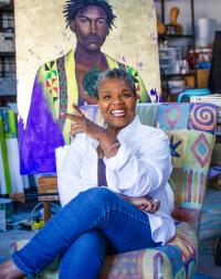 Photo of Natalie Daise.  An older Black woman sitting in a colorful patchwork chair.  A painting of a Black man is behind her.  She wears a white shirt, blue jeans, and has a smile on her face.