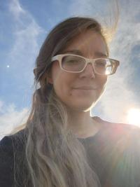 Photo of Renee Shaw.  A white woman with cream colored glasses.  She has long blondish hair tied back which spills over her right shoulder.  She is wear in a dark top.  She is in front of a blue sky with the sun beaming behind her.  