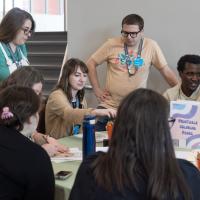 Staff members work together on a project at a table with some sitting and some standing.