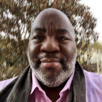Headshot of John Browne.  A black man wearing a purple collared shirt and a gray scarf.  John is bald with a graying beard.  He has a slight small and is standing in front of a slightly blurry tree.  