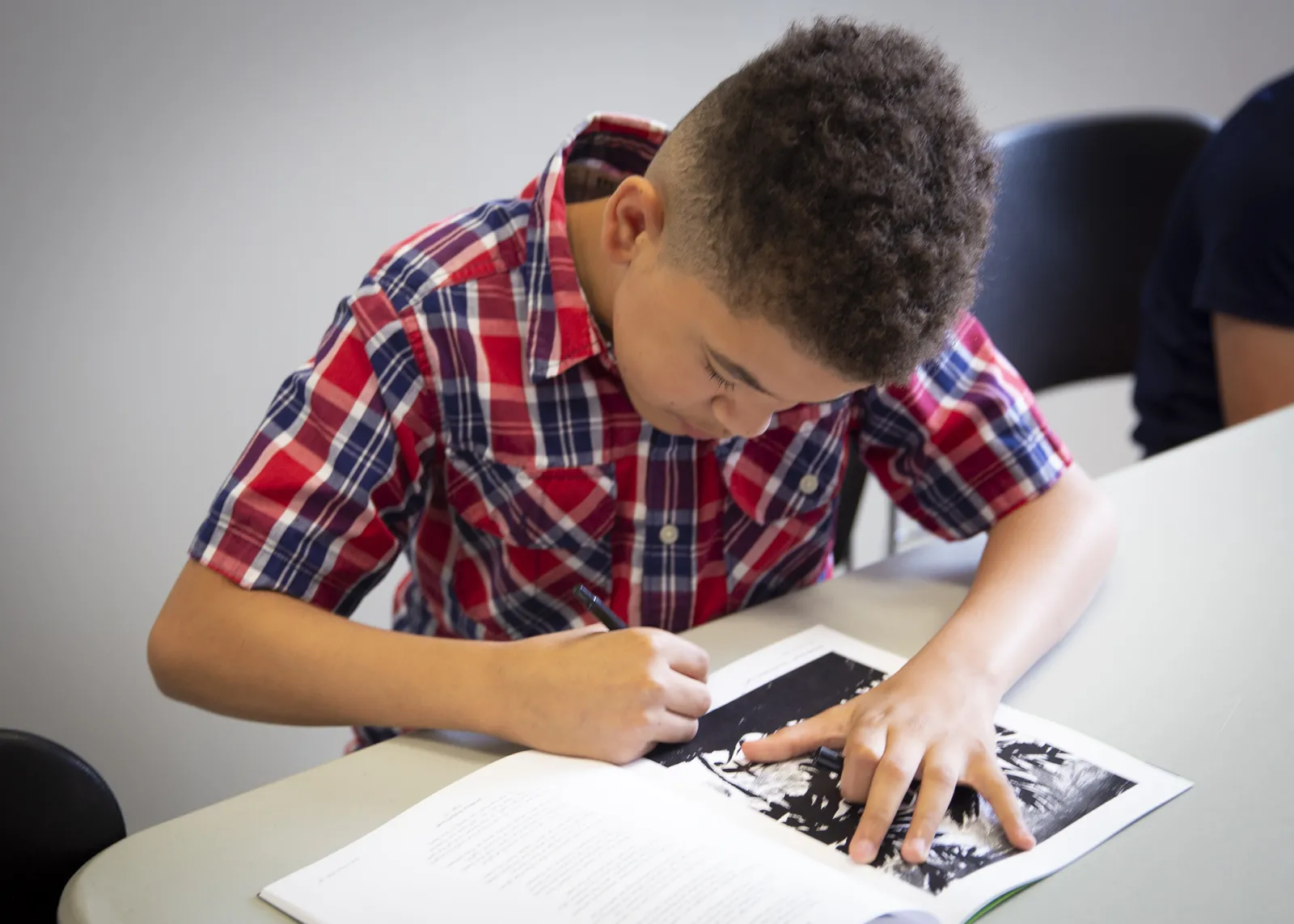 A school-aged, light-skinned Black male is bent over a copy of Kids in Print magazine and is using a black pen to sign his copy.