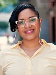Photo of Emely Rumble, a light-skinned Black woman.  She wears a light yellow shirt with a color and matching wire frame glasses with a chain.  Her head is tilted and she is smiling.  Her hair is up and her bangs are swept to the side.  