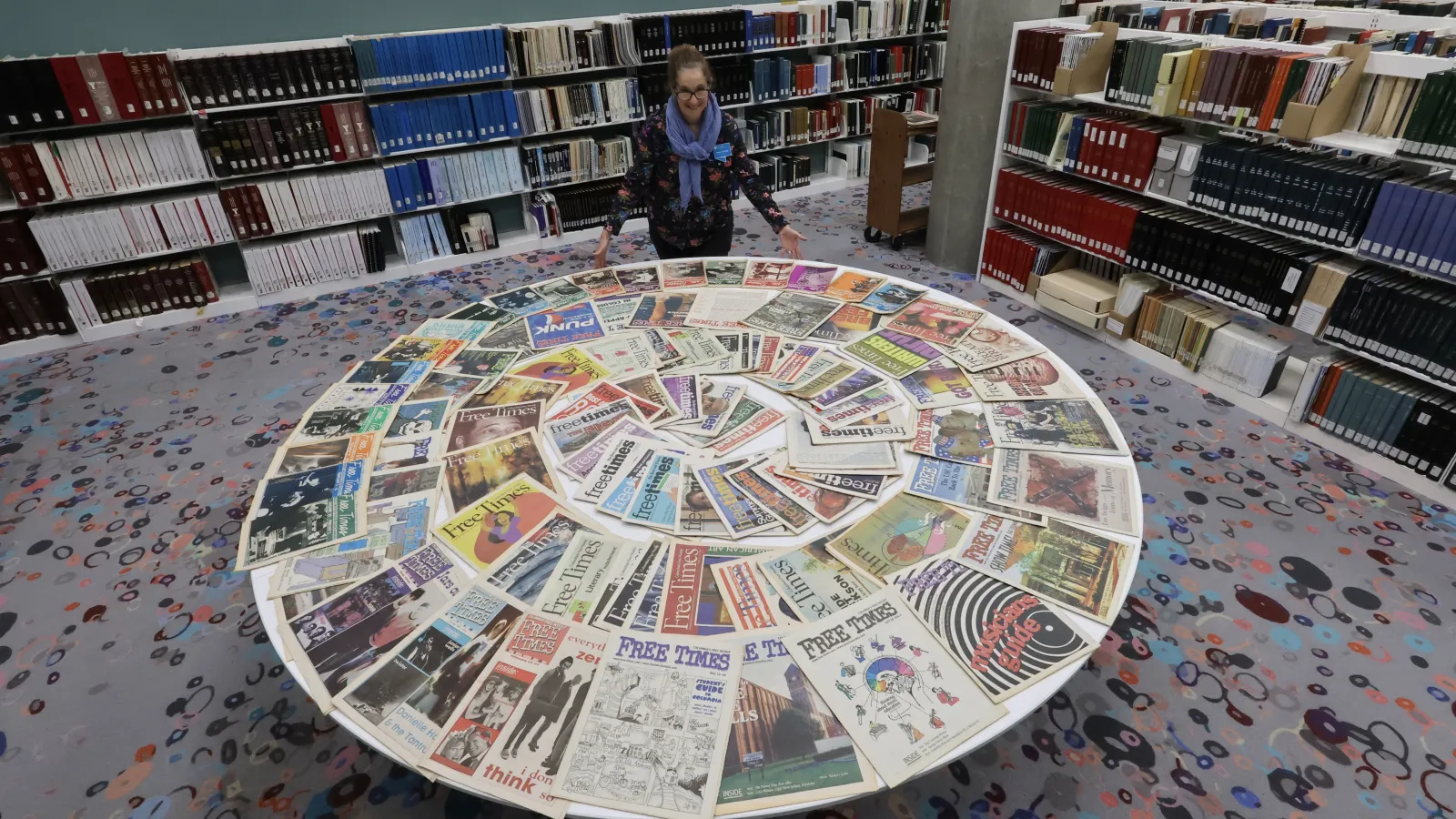A staff member in the Walker Local and Family History Center stands behind a table filled with Free Times newspaper issues arranged in a circle, highlighting the newly available archive.