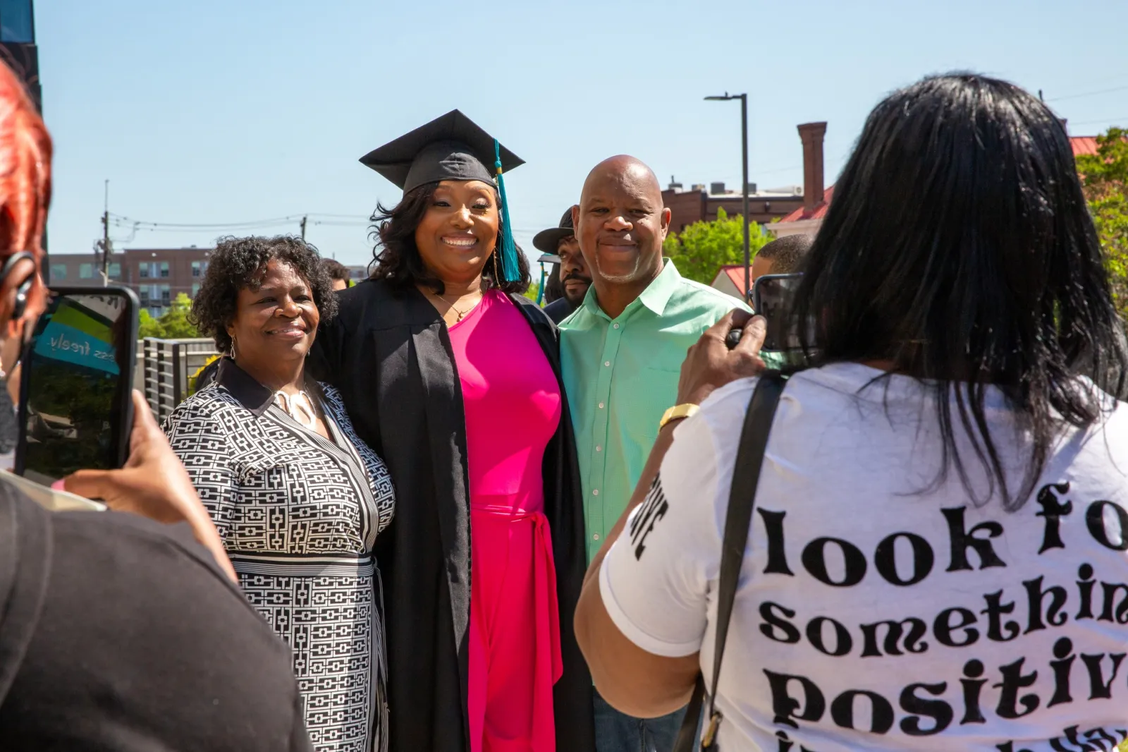 A smiling graduate in a cap and gown stands outdoors between two proud family members, posing for a photo. The graduate wears a bright pink dress under her gown. In the foreground, a woman wearing a white T-shirt that reads "look for something positive" is taking their photo with a phone. The background includes trees, a brick building, and a clear blue sky, capturing a joyful and celebratory moment.