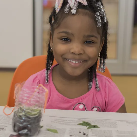 Young black female with a bright bow in her hair and a pink shirt sits in front of a table covered with newspaper during a gardening program at Richland Library