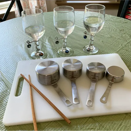 set of metal measuring cups and chopsticks on white cutting board, with three stemmed water glasses in background