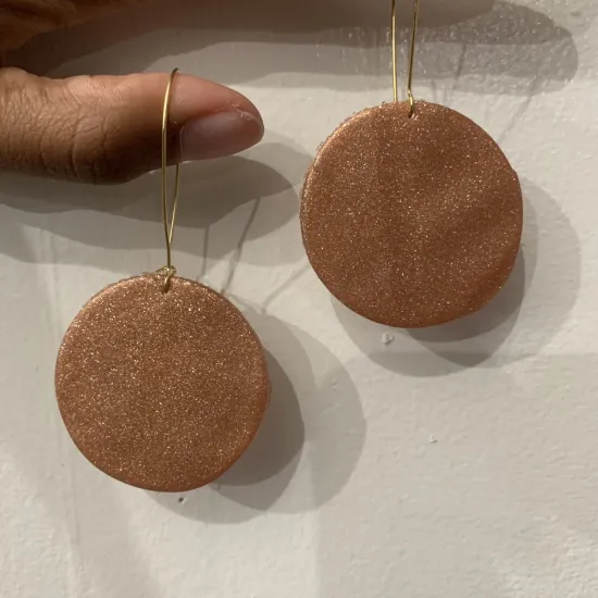 Bronze circular clay earrings with hanging thin wires hanging on two brown skinned fingers, against a white background.