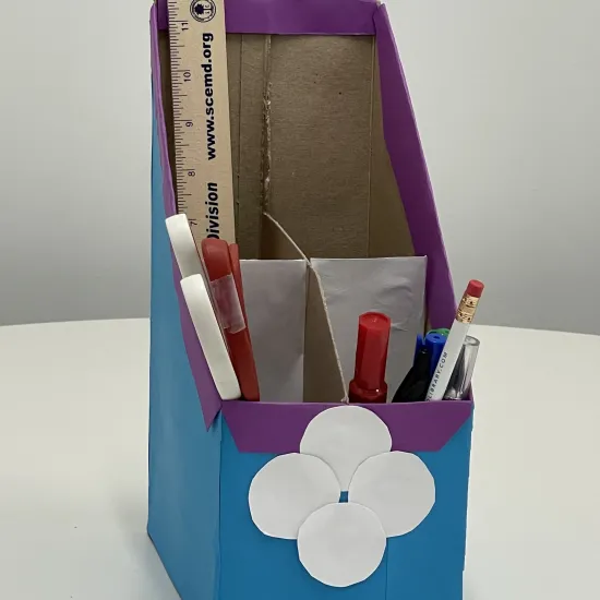 blue, purple, and white desk organizer made out of cardboard;  tan ruler, white and red scissors, red and green markers, white pencil sit are in organizer