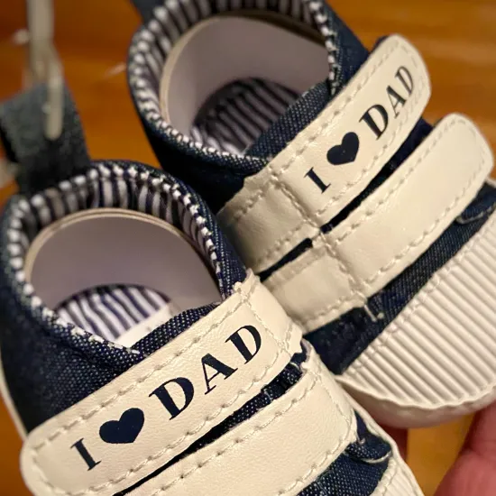A photo of a hand, holding a pair of infant shoes saying, "I love Dad"!